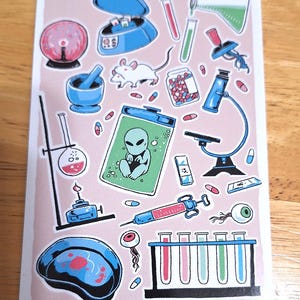May include: A sticker sheet with science-themed illustrations. Includes a microscope, test tubes, an alien, pills, a syringe, and a lab rat. The color palette is primarily pink, blue, and white, with red accents. The overall theme is laboratory and science.
