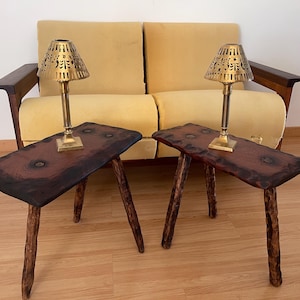 Pode incluir: Um par de mesas de apoio a condizer com tampos de madeira escura de bordo natural, nós visíveis e três pernas rústicas semelhantes a ramos. Cada mesa tem um candeeiro de latão com um suporte fino e um abajur cónico perfurado.