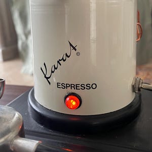 Può includere: Una base cilindrica bianca per macchina da caffè espresso con la parola Karat stampata in corsivo nero e la parola ESPRESSO in lettere stampatello nere. Una piccola spia circolare rossa è accesa sulla parte anteriore, sopra una base nera.