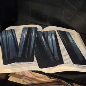 May include: Four black and white bookmarks with a forest scene. The bookmarks are laid out on a book with the text "Good to see you, Mary." I said. I'd been...