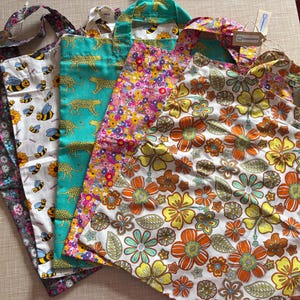 May include: A collection of five fabric tote bags featuring diverse patterns including a dark floral print, a white background with yellow and black bees, a teal green leopard print, a vibrant multi-coloured floral design, and a large orange and yellow flower print.