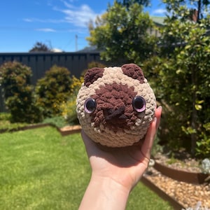 May include: A crocheted pug-shaped toy, featuring a beige and brown colour scheme. The toy has large, purple-rimmed eyes and a dark brown face and ears. The toy is held up against a backdrop of green grass and a blue sky with trees.