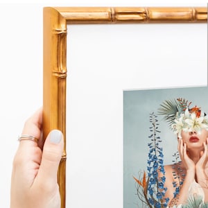 Puede incluir: Un marco de fotos dorado con una moldura texturizada estilo bambú. El marco está vacío y cuenta con un paspartú blanco en el interior. Una parte de una impresión que muestra a una persona con flores sobre los ojos es visible abajo a la derecha.