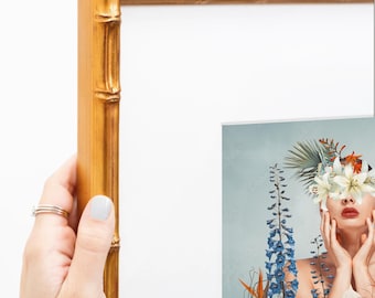 Puede incluir: Un marco de fotos dorado con una moldura texturizada estilo bambú. El marco está vacío y cuenta con un paspartú blanco en el interior. Una parte de una impresión que muestra a una persona con flores sobre los ojos es visible abajo a la derecha.