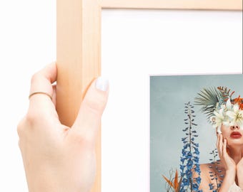 Puede incluir: Un marco de fotos de madera clara que contiene una impresión de una persona con flores cubriendo sus ojos. La persona tiene labios rojos y está rodeada de altas flores de espuela de caballero azules y hojas de palma sobre un fondo azul.