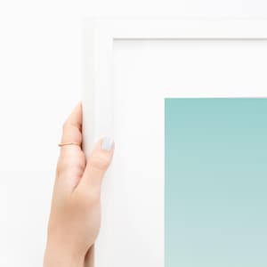 May include: A white framed artwork with a gradient of light blue to white. A hand with a gold ring and light blue nail polish holds the frame.