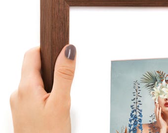 Puede incluir: Un marco de fotos rectangular con un acabado de grano de madera marrón oscuro. El marco contiene una impresión que muestra a una persona con un arreglo floral cubriendo sus ojos, rodeada de flores de espuela de caballero azules y hojas tropicales.