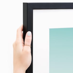 May include: A black picture frame with a white mat and a light blue and white gradient artwork. The frame is held by a hand with a gold ring and light-colored nail polish.