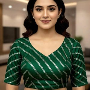 May include: A green blouse with a V-neck and short sleeves. The blouse features diagonal silver stripes across the front. The woman in the image is wearing the blouse and looking at the camera.