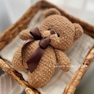 Puede incluir: Un oso de peluche marrón con un lazo de cinta marrón oscuro, descansando en una cesta tejida sobre una tela blanca. El oso tiene un aspecto texturizado y afelpado. El texto "@SHOP.HAPPY.TOYS" es visible.