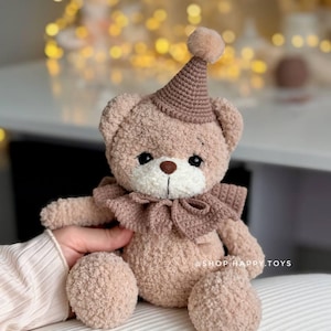 May include: A beige plush teddy bear with a textured, fuzzy finish, wearing a brown crocheted party hat with a pom-pom on top and a matching brown crocheted ruffled collar. The bear has dark eyes and a brown nose. Text reads @SHOP.HAPPY.TOYS.