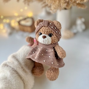 May include: A brown crocheted teddy bear wearing a brown crocheted dress with white pearl accents. The bear has brown eyes and a brown nose.