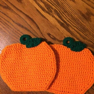 Puede incluir: Dos agarraderas de olla de calabaza naranja tejidas a crochet con tallos verdes.