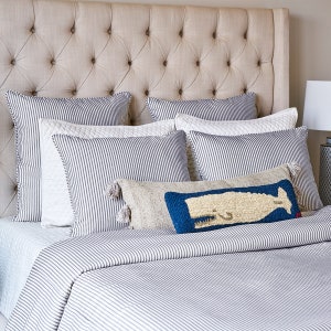 Puede incluir: Una cama con cabecero capiton&eacute; beige, ropa de cama a rayas blancas y azul marino, y varios cojines decorativos. Un coj&iacute;n rectangular presenta un dise&ntilde;o de ballena texturizado en azul y crema con borlas en los extremos al frente.