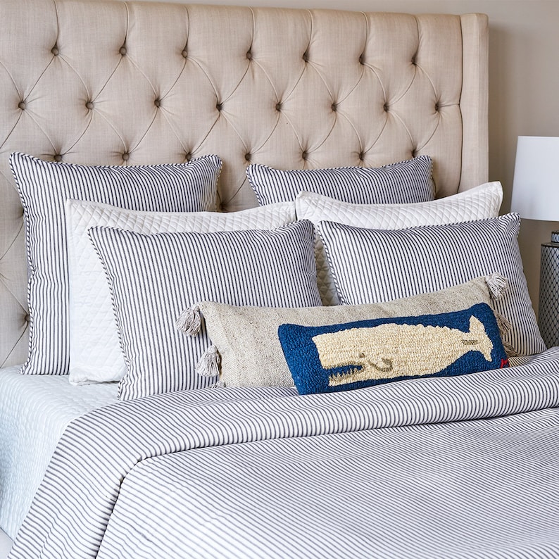 Puede incluir: Una cama con cabecero capiton&eacute; beige, ropa de cama a rayas blancas y azul marino, y varios cojines decorativos. Un coj&iacute;n rectangular presenta un dise&ntilde;o de ballena texturizado en azul y crema con borlas en los extremos al frente.