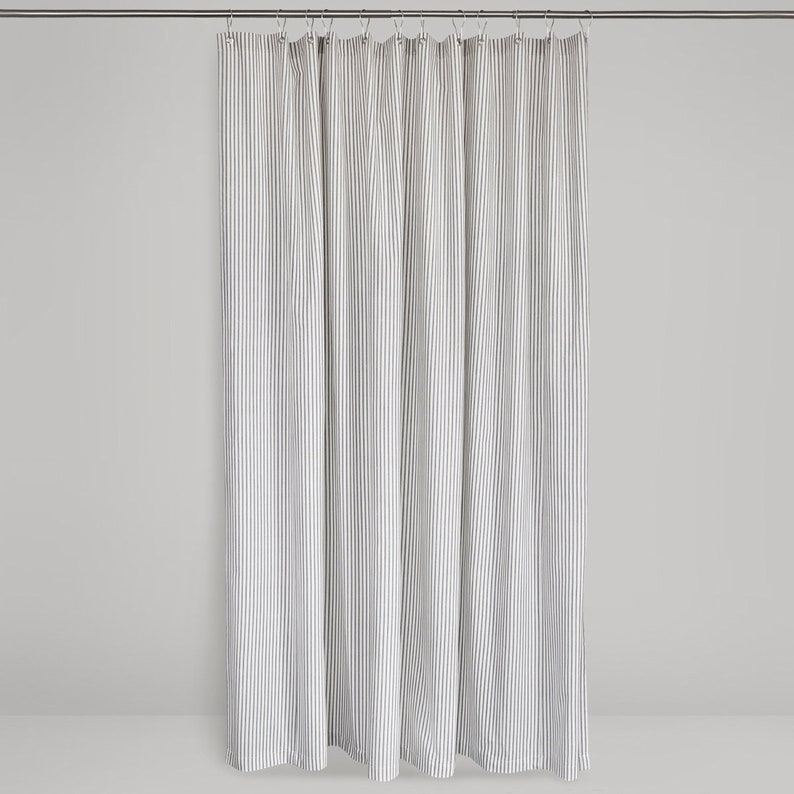 image featuring a gray and white fabric shower curtain in gray and cream ticking stripe fabric hanging on a shower curtain rod against a neutral background