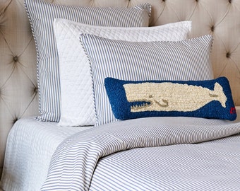 Puede incluir: Un conjunto de ropa de cama que incluye una funda nórdica de rayas azules y blancas, dos almohadas a rayas a juego, una almohada blanca acolchada y un cojín rectangular con una ballena color crema sobre fondo azul oscuro.
