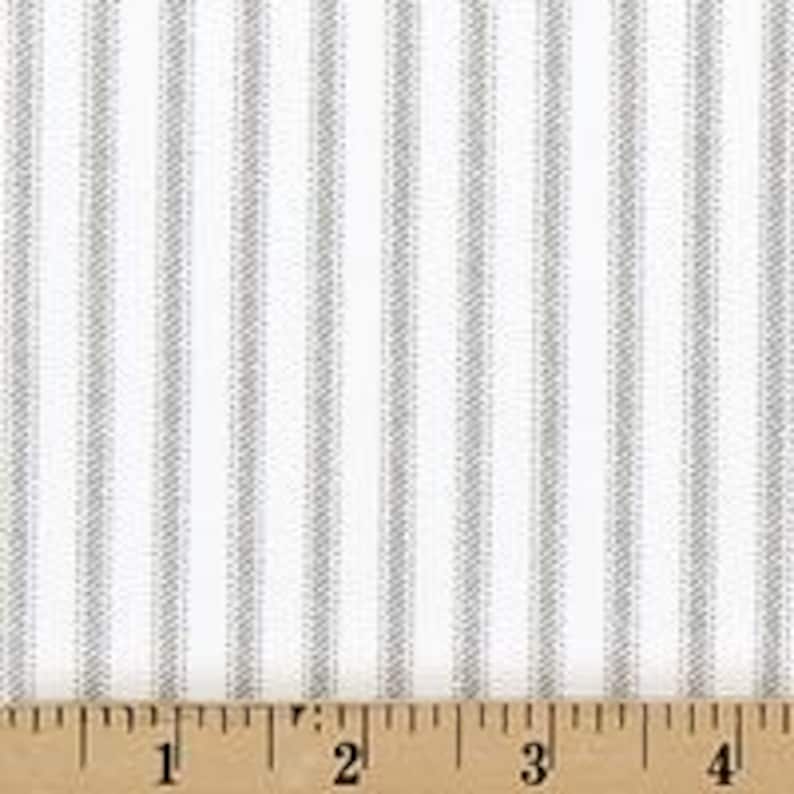 Puede incluir: Una tela con un patr&oacute;n repetitivo de finas rayas verticales grises sobre un fondo blanco s&oacute;lido. Una regla de madera est&aacute; colocada en el borde inferior, mostrando una escala en pulgadas que va de cero a cuatro pulgadas a lo ancho de la tela.