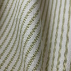 Puede incluir: Una tela tejida con un patr&oacute;n repetitivo de rayas verticales de color verde salvia sobre un fondo blanco. El material muestra una textura de tejido visible con pliegues suaves que crean profundidad y dimensi&oacute;n en toda la superficie.