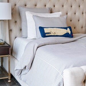 Puede incluir: Una cama con funda n&oacute;rdica de rayas azules y blancas y fundas de almohada a juego. Una almohada blanca acolchada se sit&uacute;a tras un coj&iacute;n rectangular azul con un gr&aacute;fico de ballena beige texturizado y un peque&ntilde;o detalle rojo en la esquina.