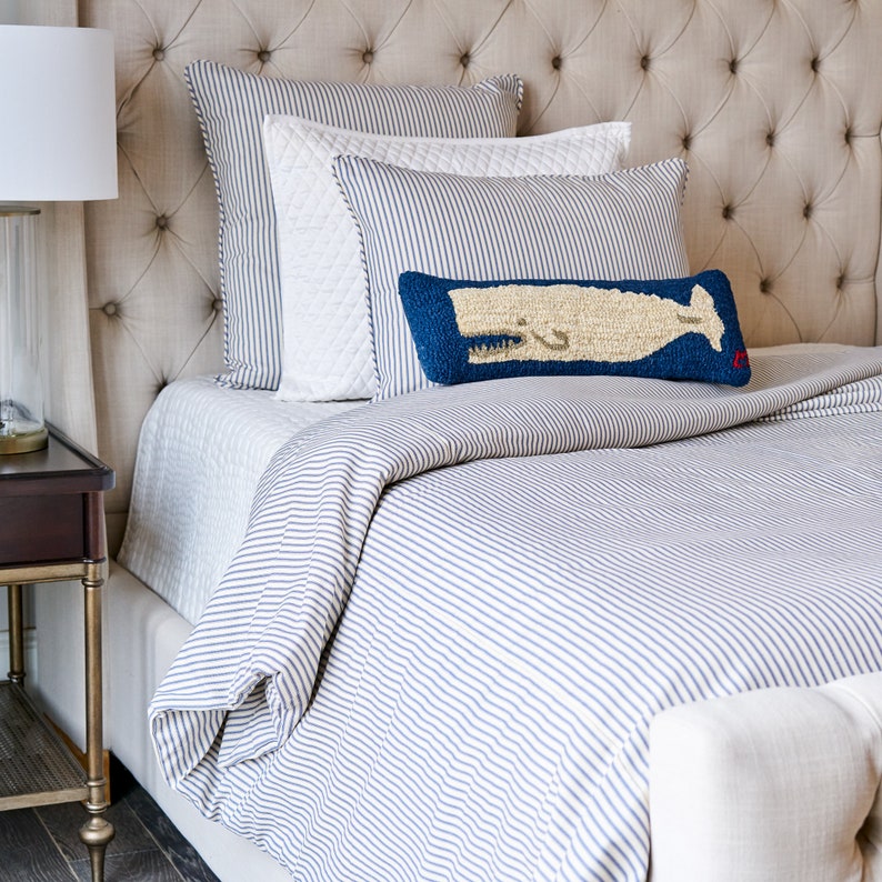 Puede incluir: Una cama con funda n&oacute;rdica de rayas azules y blancas y fundas de almohada a juego. Una almohada blanca acolchada se sit&uacute;a tras un coj&iacute;n rectangular azul con un gr&aacute;fico de ballena beige texturizado y un peque&ntilde;o detalle rojo en la esquina.