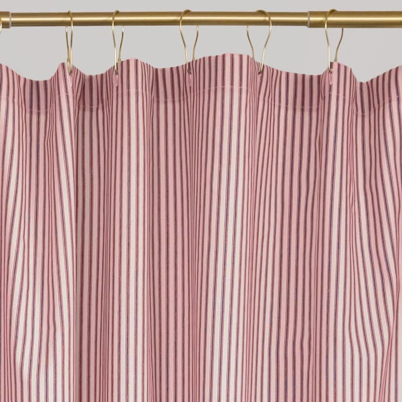 Puede incluir: Una cortina de ducha con un patr&oacute;n de rayas verticales en tonos rosa claro, blanco y azul oscuro. La tela cuelga de una barra de metal dorado mediante ganchos de metal dorado a juego, espaciados uniformemente a lo largo del borde superior.