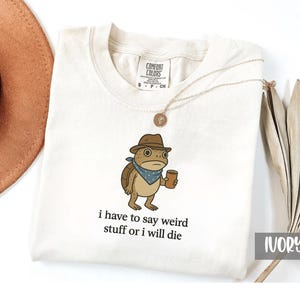 May include: Ivory-colored Comfort Colors t-shirt featuring a cartoon turtle wearing a hat and holding a cup. The text "i have to say weird stuff or i will die" is printed below the turtle graphic. A gold necklace and a brown hat are also visible.