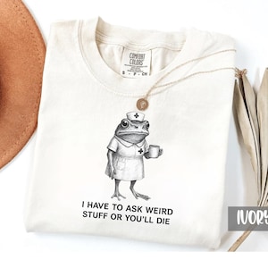 May include: Ivory-colored t-shirt with a nurse frog graphic and the text "I HAVE TO ASK WEIRD STUFF OR YOU'LL DIE." The shirt features a crew neck and the Comfort Colors brand label. A gold necklace with a pendant is draped on the shirt.