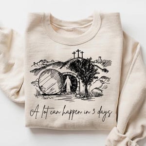 Op de afbeelding: Een crèmekleurige sweatshirt met een zwarte afbeelding van een religieuze scène, waaronder drie kruisen op een heuvel, een boom en een figuur die uit een graf komt. De tekst "A lot can happen in 3 days" staat eronder.