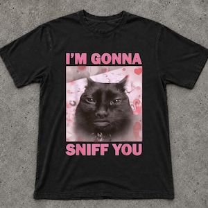 Pode incluir: Uma t-shirt preta de manga curta com um gráfico central de um gato preto sobre um fundo cor-de-rosa com padrões de corações. O texto "I'M GONNA" aparece em cor-de-rosa acima do gato, e "SNIFF YOU" está impresso abaixo da imagem.