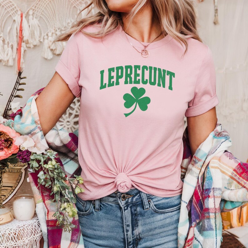May include: A light pink t-shirt with the word "LEPRECONT" in green block letters and a green shamrock. The shirt is knotted at the waist and paired with blue jeans. A plaid shirt is draped over the shoulders.