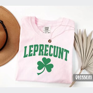 May include: A light pink t-shirt with the word "LEPRECONT" in green block letters and a green shamrock. The shirt has a crew neck and short sleeves. The brand name "COMFORT COLORS" is printed on the tag. A gold necklace is on the shirt.