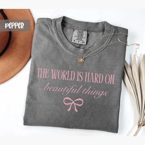 May include: A gray Comfort Colors t-shirt with the text "THE WORLD IS HARD ON beautiful things" in pink, along with a pink bow graphic. A silver necklace with a pendant rests on the shirt. A brown hat is visible in the background.
