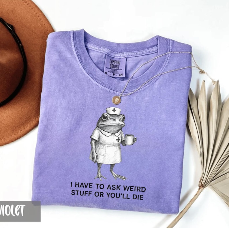Op de afbeelding: Een violet Comfort Colors t-shirt met een afbeelding van een kikker verkleed als verpleegster, met een mok. De tekst "I HAVE TO ASK WEIRD STUFF OR YOU'LL DIE" staat eronder. Een gouden ketting en een bruine hoed zijn ook zichtbaar.