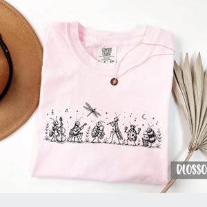 May include: A light pink t-shirt featuring a black ink illustration of various insects playing musical instruments, including an ant with a bass, a cricket with a guitar, a beetle with a saxophone, a mantis with a flute, and a bee with a drum.