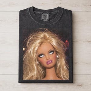 Può includere: Una t-shirt grigio antracite a girocollo con una grande stampa grafica di una bambola con lunghi capelli biondi, trucco marcato e orecchini di perle. La bambola ha lo sguardo rivolto verso l'alto e una bocca dai toni rosati sul davanti.