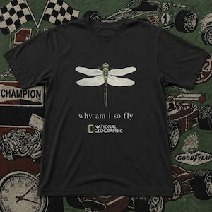 以下が含まれることがあります： 胸の中央にトンボのグラフィックがプリントされた黒の半袖Tシャツ。トンボの下には「why am i so fly」というテキストがあり、その下に黄色い長方形の中に白い文字でNational Geographicのロゴが配置されています。