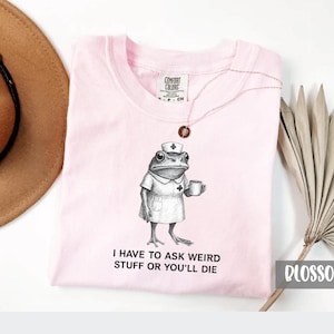 Op de afbeelding: Lichtroze Comfort Colors T-shirt met een illustratie van een verpleegster-kikker die een mok vasthoudt. De tekst "I HAVE TO ASK WEIRD STUFF OR YOU'LL DIE" staat eronder. Een ketting met een hanger is zichtbaar.