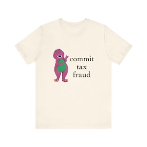 May include: A cream-coloured t-shirt with a purple dinosaur character with a green belly, gesturing with its hand. The text "commit tax fraud" is printed in black. The shirt is made of a soft material.