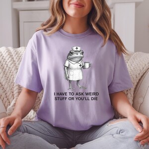 Op de afbeelding: Een lichtpaars T-shirt met een grappige illustratie van een kikker in een verpleegstersuniform, met een mok. De tekst "I HAVE TO ASK WEIRD STUFF OR YOU'LL DIE" staat eronder. Het shirt is een casual unisex stijl.