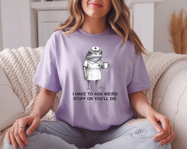 Op de afbeelding: Een lichtpaars T-shirt met een grappige illustratie van een kikker in een verpleegstersuniform, met een mok. De tekst "I HAVE TO ASK WEIRD STUFF OR YOU'LL DIE" staat eronder. Het shirt is een casual unisex stijl.