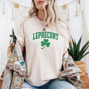 May include: A cream-colored t-shirt with the word "LEPRECONT" in green block letters and a green shamrock graphic. The shirt is part of a St. Patrick's Day theme and is made of a soft material.