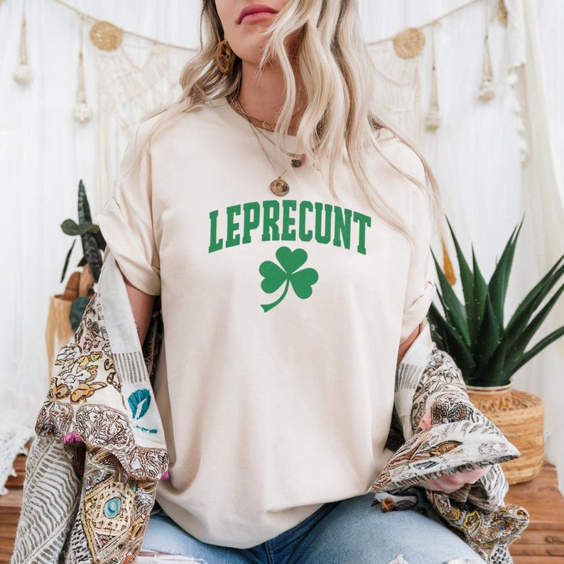 May include: A cream-colored t-shirt with the word "LEPRECONT" in green block letters and a green shamrock graphic. The shirt is part of a St. Patrick's Day theme and is made of a soft material.