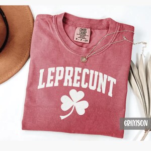 May include: Crimson-colored t-shirt with the word "LEPRECUNT" in white block letters and a white shamrock graphic. The shirt has a crew neck and short sleeves. A silver necklace with a pendant is draped across the shirt.