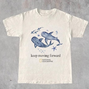 Pode incluir: Uma t-shirt de manga curta cor de creme com uma ilustração em aguarela azul de dois tubarões, uma estrela-do-mar e peixes pequenos. Abaixo do gráfico, o texto "keep moving forward" e o logótipo da National Geographic em preto.