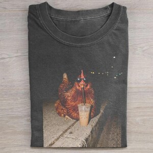 Könnte beinhalten: Ein anthrazitfarbenes T-Shirt mit Rundhalsausschnitt und einem Grafikdruck eines braunen Huhns, das eine schwarze Sonnenbrille trägt und aus einem durchsichtigen Plastikbecher mit Strohhalm auf einer Betonoberfläche trinkt.