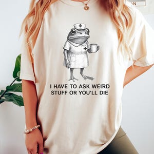 Op de afbeelding: Cr&egrave;mekleurig T-shirt met een grillige illustratie van een kikker verkleed als verpleegster, met een mok. De tekst "I HAVE TO ASK WEIRD STUFF OR YOU'LL DIE" staat eronder. Het T-shirt wordt door een persoon gedragen.
