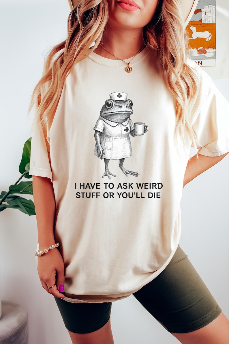 Op de afbeelding: Cr&egrave;mekleurig T-shirt met een grillige illustratie van een kikker verkleed als verpleegster, met een mok. De tekst "I HAVE TO ASK WEIRD STUFF OR YOU'LL DIE" staat eronder. Het T-shirt wordt door een persoon gedragen.