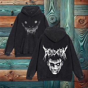May include: Black hoodie with a front pocket and a hood. The front features a white, stylized grinning face. The back displays the word "BERSERK" above a white illustration of a face with dark markings. The hoodie is made of a soft, comfortable material.