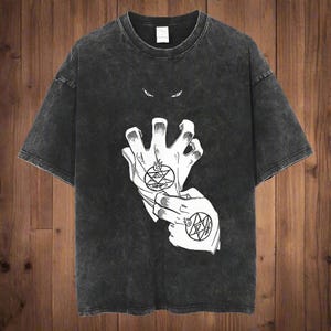 May include: Dark gray t-shirt featuring a white graphic of two hands with alchemic symbols and a pair of eyes above. The shirt has a crew neck and short sleeves. The fabric has a washed, vintage look.